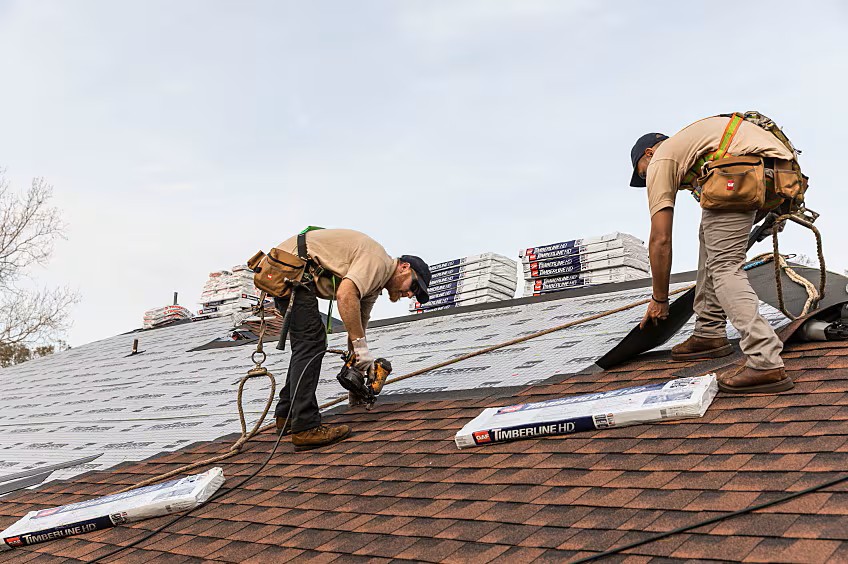 Two roofing installers fastening GAF Timberline HD shingles with branded packages staged on the roof